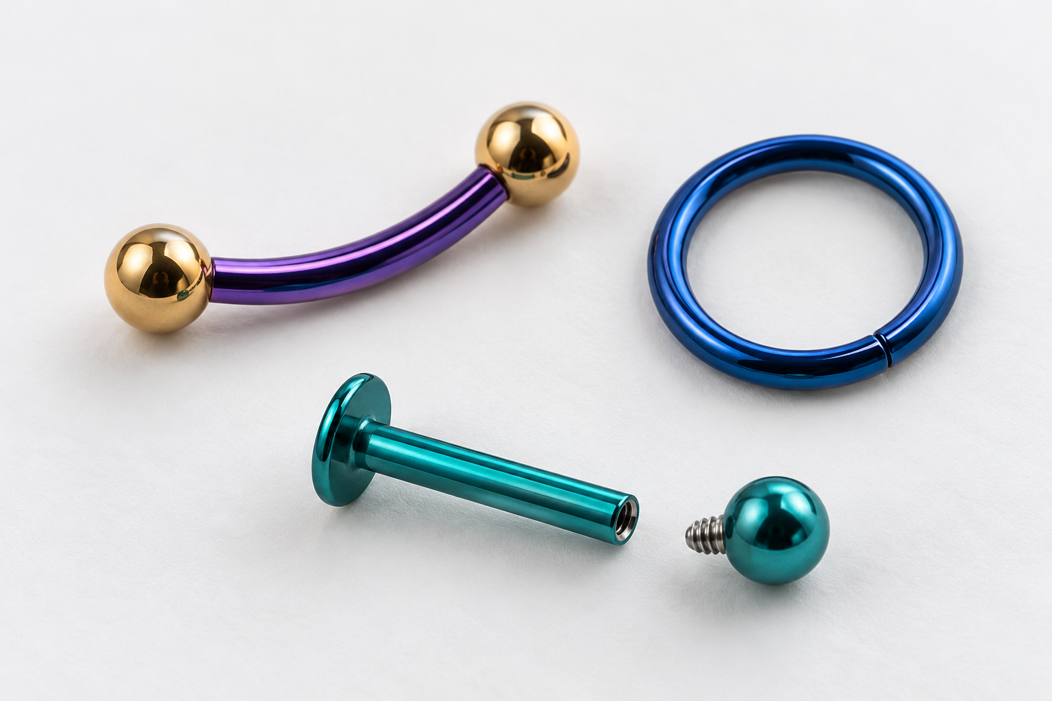 Três joias de piercing sobre fundo branco: uma barra curva roxa, um anel azul e uma joia labret turquesa com esfera separada.