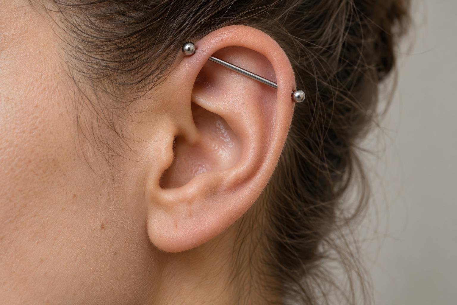 Orelha adulta com piercing Industrial de barra reta, fotografada de perfil para mostrar a relação da joia com o bordo e a concavidade da orelha.